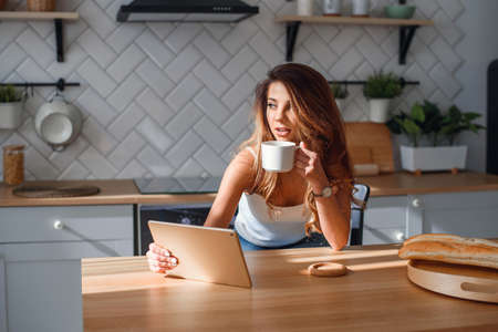Pretty young woman using tablet while drinking morning coffee after breakfast on the kitchen.の写真素材
