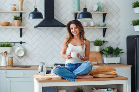 Pretty happy woman drinks coffee while sitting on the table on the cozy kitchen in the morning at home. Morning relaxation at home.の写真素材