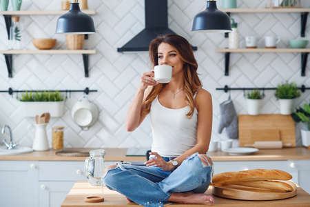 Pretty happy woman drinks coffee while sitting on the table on the cozy kitchen in the morning at home. Morning relaxation at home.の写真素材