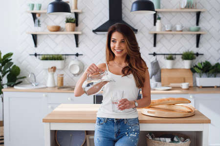 Smiling pretty woman pours pure water from the bottle into the glass in the kitchen. Healthcare and diet concept.の写真素材