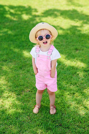 A smiling little girl in pink sunglasses holds her arms in her pockets and looks at the camera. Summer vacation concept.の写真素材