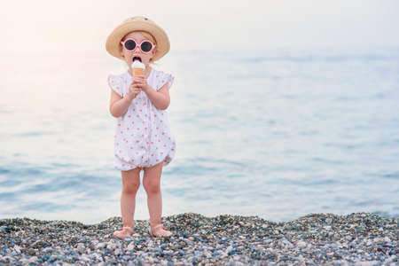 Funny toddler girl wearing pink summer overalls, hat and pink sunglasses eats vanilla white ice cream on the vacations on the sea beach.の写真素材