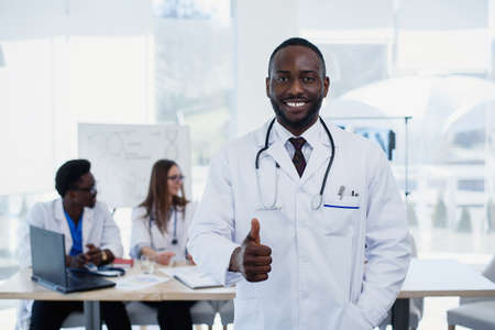 Portrait of smiling afro american male doctor showing thumb up sign. Medical assistant or student in white uniform with stethoscope. Handsome male doctor with a medical group at meeting room.の写真素材