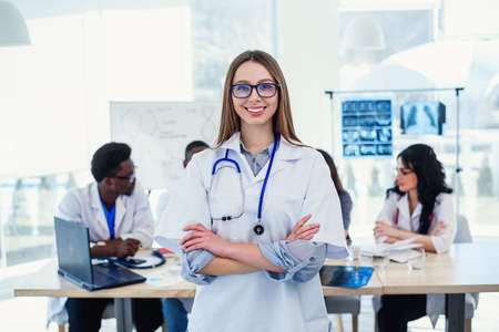 Smiling medical doctor woman with stethoscope standing in front of medic team at hospital. Attractive young female doctor in white coat and glasses looking at the camera.の写真素材