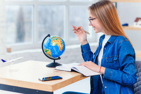 Young caucasian woman sits at the table and rotates the globe to choose the country for vacation and...