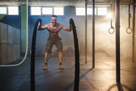 Handsome strong man doing exercises for the muscles with battle ropes in fitness gym.