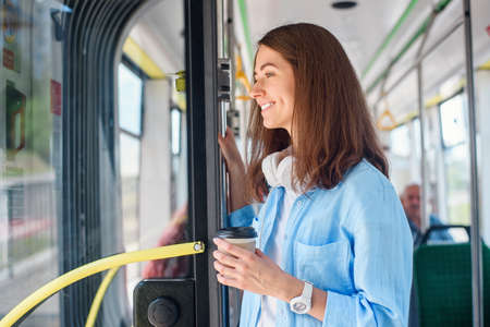 Stylish woman in blue shirt enjoying trip in the modern tram or bus, stands with cup of coffee in the public transport.の写真素材