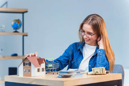Young pretty girl counts her savings money and looks to a model of the house.の写真素材