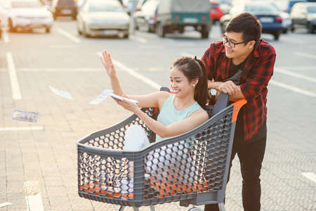 Lovely cheerful vietnamese girl scattering money ,sitting in the shopping trolley which holding her attractive boyfriendの写真素材