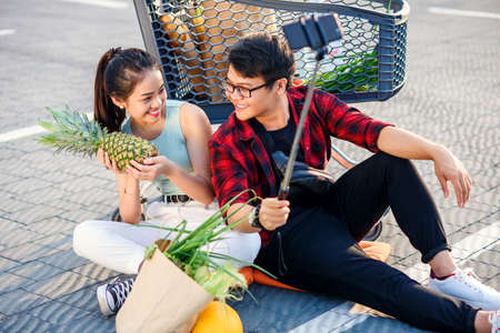 Happy vietnamese couple sitting on the ground near big shopping centre and making selfie photo with peanapple.の写真素材