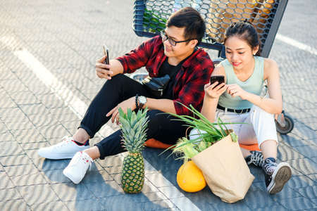 Attractive vietnamese boy and beauiful girl sitting on the parking near shopping mall and uses their smartphones.の写真素材