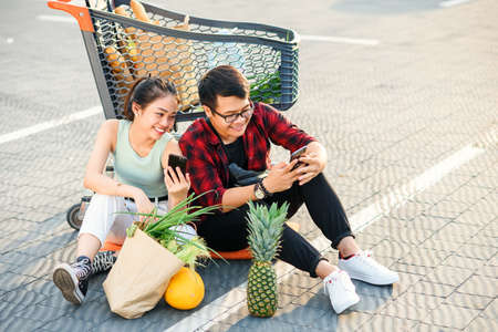 Attractive vietnamese boy and beauiful girl sitting on the parking near shopping mall and uses their smartphones.の写真素材
