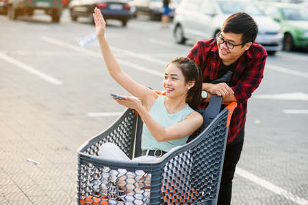 Lovely cheerful vietnamese girl scattering money ,sitting in the shopping trolley which holding her attractive boyfriendの写真素材