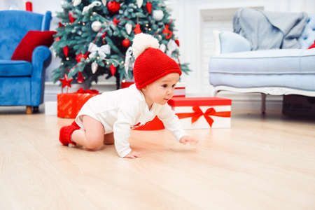 Adorable smiling infant crawling on the parquet to the christmas gifts near his mom which sits on...