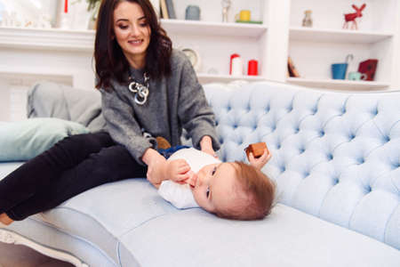 Cute toddler lies on the blue couch and happy young mommy kneading his legs and makes him happy.