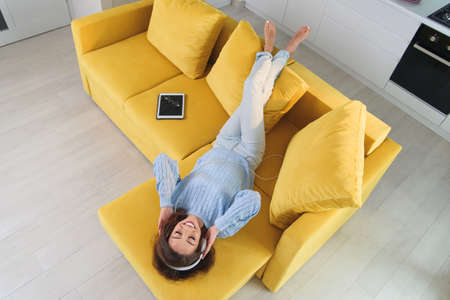 Satisfied cheerful girl lying on the comfortable yellow couch and listens to the music on her white earphones. Top view.の写真素材