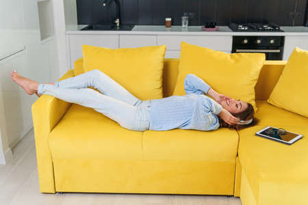 Pretty teenage girl in headphones listening to music while lying on her back on sofa at modern stylish kitchen. Young happy woman relaxing at cozy home.の写真素材