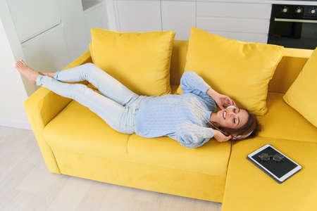 Pretty teenage girl in headphones listening to music while lying on her back on sofa at modern stylish kitchen. Young happy woman relaxing at cozy home.の写真素材