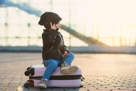 Cute little traveler girl sits on pink suitcase and talking on smartphone near airport against sunset. Modern high developed young generation using gadgets.の写真素材