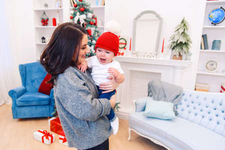 Happy beautiful mother having fun with her infant son near Christmas Tree in Christmas evening.