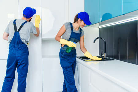 Attractive young couple of man and woman in blue uniform cleaning the furniture with wipes and spray in the comfortable cuisineの写真素材