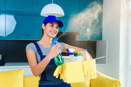 Cheerful girl in special uniform and yellow rubber gloves holds cleanser spray with cleaning fluid.