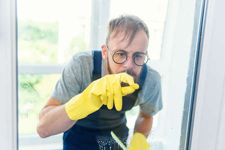 Cheerful bearded man in funny eyeglasses cleans the window with detergents and remove dirt with his finger in gloves.の写真素材