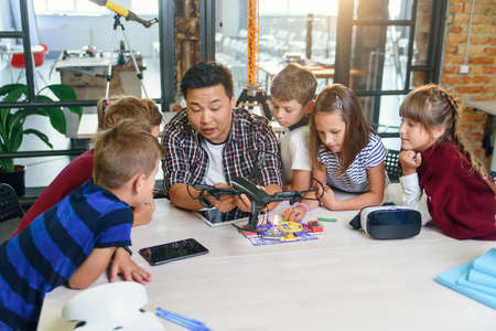 In physics and mechanics lesson young Asian teacher demonstrates quadcopter for caucasian pupils at class in modern smart school. Science, drone, engineering and future concept.の写真素材