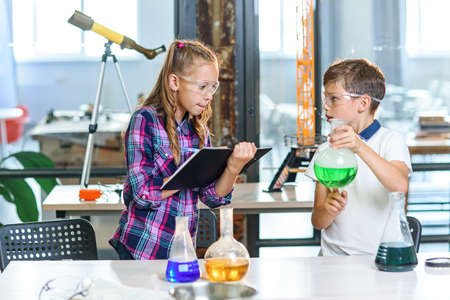 Children record results of the experiments in notebook. Two young clever caucasian pupils in protective glasses doing experiment with green liquid in beaker and dry ice.の写真素材