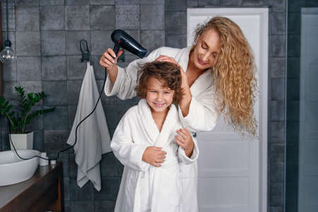 Beautiful mom dries hair with a hairdryer to her little happy son, dressed in a white bathrobe.の写真素材