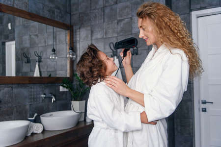 Beautiful mom dries hair with a hairdryer to her little happy son, dressed in a white bathrobe.の写真素材