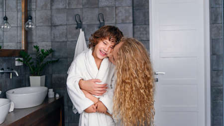 Beautiful mother with curly hair in a white bathrobe hugs her little smiling son in the bathroom.の写真素材