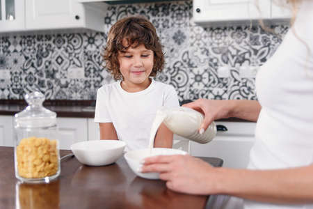 Beautiful mother pours milk into a plate with cereal Beautiful mother prepares breakfast for her...