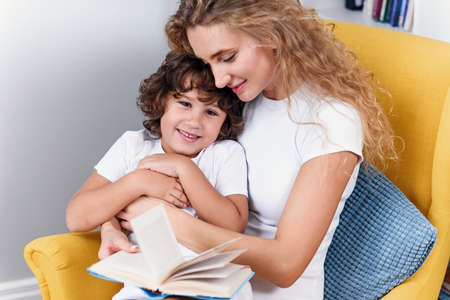 Beautiful mother reads child book to her little son while sitting in comfortable yellow chair near window at cozy home.の写真素材