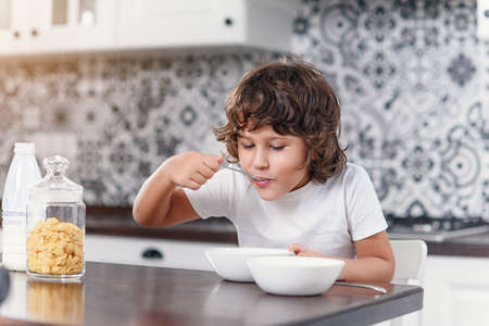 Happy little boy eats with pleasure healthy breakfast of cornflakes and milk at the kitchen.の写真素材