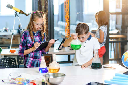 Children record results of the experiments in notebook. Two young clever caucasian pupils in protective glasses doing experiment with green liquid in beaker and dry ice.の写真素材