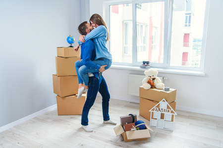 Pretty smiling girl which holds by her handsome young boyfriend in the middle of their new flat full...