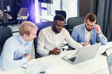 Three businessmen sitting at the modern office tables and analysing data from report and computer to succeed in developing of their own company.の写真素材