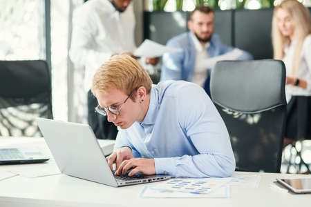 Cheerful purposeful male office worker with funny eyeglasses sits at his workplace and using laptop...