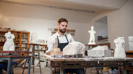 Male sculptor in white shirt and black apron makes a limestone copy of woman torso at the artistic...