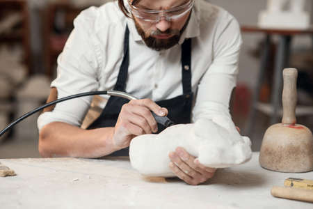 Attentive stonemason with stylish beard in protective goggles detailing creative sculpture of woman torso at his workplace.の写真素材