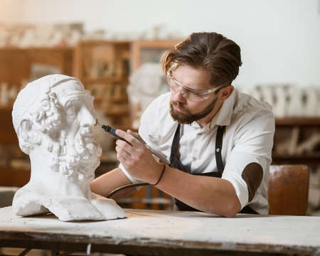 Professional bearded carver polishing with micro drill machine details of the mans sculpture head in...