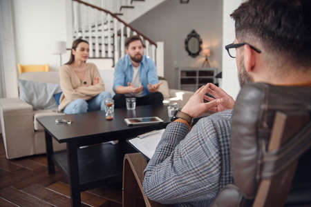 Close up of male psychologist sits at chair and listens depressed young couple at stylish cozy office. Selective focus.の写真素材