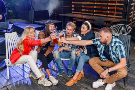 Great time with friends. Four young cheerful people chatting and drinking beer while sitting at the chairs on the yard back house on the weekend evening. Outdoors. Slow motionの写真素材