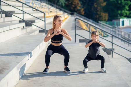Sporty beautiful elder and younger sisters which squating together during fitness training outdoors.