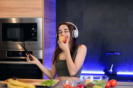 Young attractive woman eats red apple and listens to the music at kitchen in morning. Healthy...