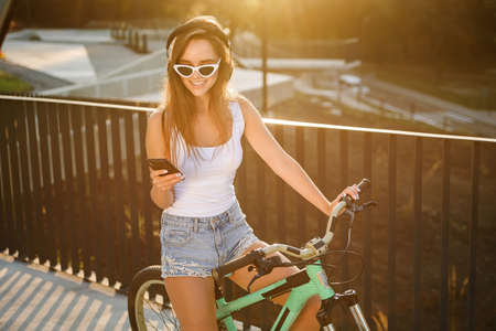 Charming sexy girl in sunglasses and tempting clothes sits on bicycle and listens to music with earphones on the sunset background. Enjoying beautiful life.の写真素材