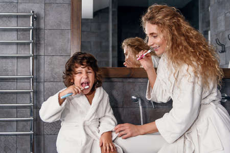 Attractive smiling young mother in dressing gown cleans her teeth together with handsome little son near washbasin with mirror. Happy family concept.の写真素材