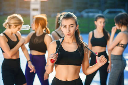 Pretty young woman with jump rope on her neck looking at camera after outdoors workout with her female friends on the background on the stadium.の写真素材