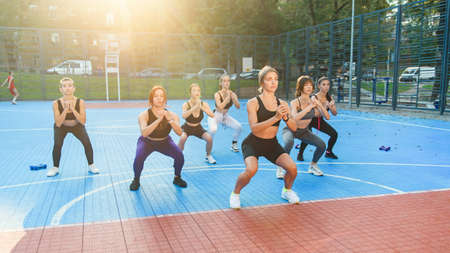Group of attractive young athletic girls in sportswear doing exercises with squats on the sportground in the green parkの写真素材